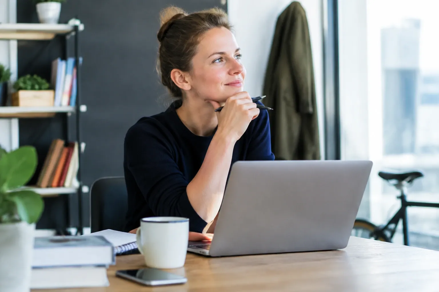 Vrouw achter laptop kijkt dromerig uit raam, pen in hand, koffiemok naast haar